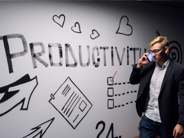 Photo of a middle-aged man in a suit holding a cell phone and standing in front of a sign with the word productivity and graphics like arrows, hearts, pencils, and notes to illustrate toxic productivity.